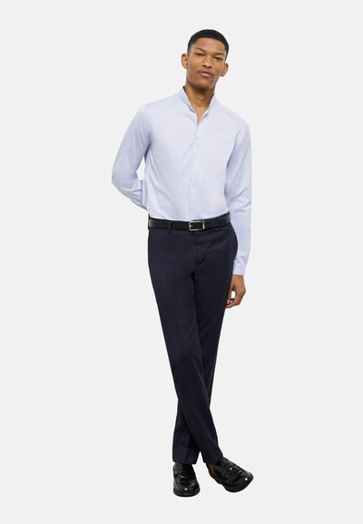 Camicia azzurra chiara con colletto alla coreana, abbinata a pantaloni blu marina scuri. Il modello indossa mocassini neri. Design semplice e su misura.