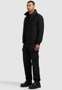 Svart bomberjakke med høy krage, glidelåslukking, sidelommer og ribbestrikkede mansjetter, kombinert med svarte cargobukser og klumpete svarte støvler.