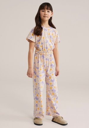 Fată tânără în picioare, purtând un combinezon cu mâneci scurte cu modele de soare și frunze portocalii și violete, asortat cu pantofi slip-on bej.