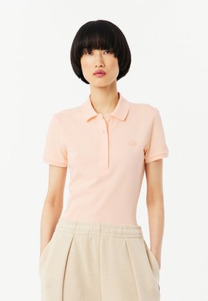 Femme aux cheveux courts noirs portant un polo rose pâle et un pantalon plissé beige taille haute, debout les mains dans les poches.