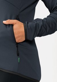 Giacca atletica grigio scuro con superficie testurizzata, tasca con zip e dettagli in nero e verde a contrasto lungo l'orlo e la zip.