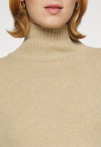 Femme portant un pull à col roulé côtelé beige et des boucles d’oreilles anneaux dorés, avec des cheveux courts châtain clair et des lèvres naturellement roses.