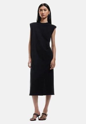 Schwarzes ärmelloses Kleid mit lockerer Passform, knielang, glatter Textur, rundem Ausschnitt und seitlichen Schlitzen, kombiniert mit schwarzen Riemchensandalen.