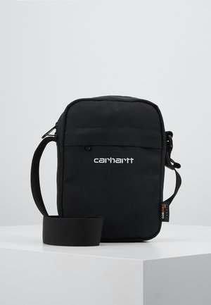 Kleine schwarze Carhartt Crossbody-Tasche mit verstellbarem Riemen, Frontreißverschlusstasche und Cordura-Stoffetikett an der Seite, auf weißer Oberfläche liegend.