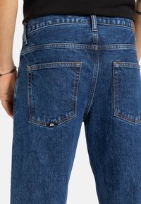 Modré džíny z denimu so strihom typu straight-cut, s dvoma zadnými vreckami a kontrastným oranžovým prešívaním. Minimalistické značkovanie na vrecku.