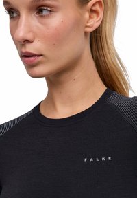 Zbliżenie na kobietę o jasnej karnacji i blond włosach, ubraną w czarną bluzkę z okrągłym dekoltem z logo "FALKE" oraz paskami na ramionach.