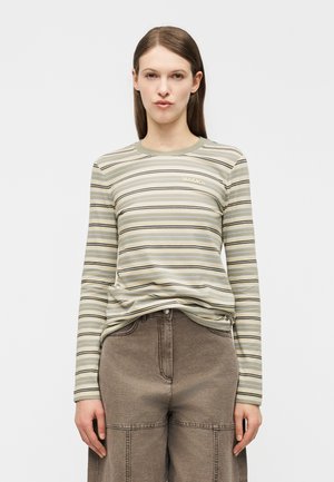 Žena nosi dlhý rukáv s béžovými, zelenými a čiernymi pruhmi, zastrčený do hnedých vysokých nohavíc na čistom bielom pozadí.