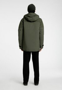 Olivgrön, fodrad jacka med huva och en slät yta, med långa ärmar och sidofickor, i kombination med svarta byxor och en stickad mössa.