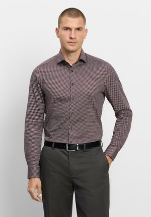 Uomo che indossa una camicia a maniche lunghe con motivo infilata in pantaloni grigio scuro con cintura nera, in piedi davanti a uno sfondo chiaro e uniforme.