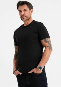 Maglietta nera a maniche corte in cotone, con scollatura rotonda, forma aderente, abbinata a jeans in denim scuro, con tatuaggi visibili sulle braccia.