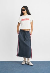 Κομμένο λευκό T-shirt με κόκκινο γραφικό "Air Jordan Fight Club", συνδυασμένο με μπλε maxi φούστα που έχει κόκκινες και λευκές πλευρικές λωρίδες. Ορατά αθλητικά παπούτσια.