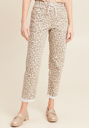 Femme portant un pantalon beige et blanc à imprimé léopard avec des ourlets retroussés et des mocassins beiges, debout devant un arrière-plan neutre.