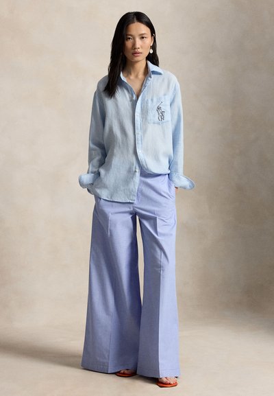 Γυναίκα που φοράει ανοιχτό μπλε, χαλαρή πουκάμισο με κουμπιά και λογότυπο παίκτη πόλο, συνδυασμένη με φαρδιά λιλά παντελόνια, στέκεται με τα χέρια στις τσέπες μπροστά σε ουδέτερο φόντο.