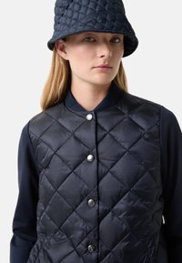 Marineblaues gesteppte Weste mit rundem Kragen, silbernen Knopf-Akzenten und glänzendem Finish, kombiniert mit einem passenden Bucket Hat.