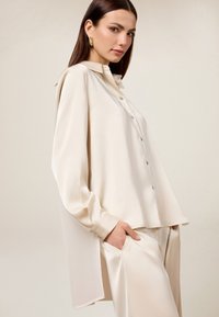 Blusa de satén color crema con mangas largas, cierre frontal de botones y un corte ligeramente oversized. Presenta un cuello clásico y aberturas laterales.