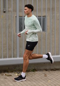 Light mint long-sleeve athletic shirt, black compression shorts, black running shoes with orange accents, white ankle socks, running posture.