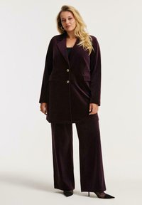 Paarse fluwelen pak met een lange blazer met twee knopen, zijzakken en brede broekspijpen. Gecombineerd met zwarte puntige hakken.