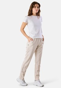 Beige joggerbyxor med sidofickor, elastisk midja och en logotypaccent på benet, i kombination med en enkel vit t-shirt och vita sneakers.