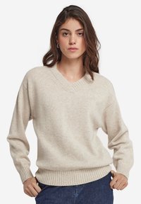 Beige v-ringad tröja i mjukt material, med ribbade manschetter och nederkant, samt en avslappnad passform. Bärs tillsammans med mörkblå jeans.