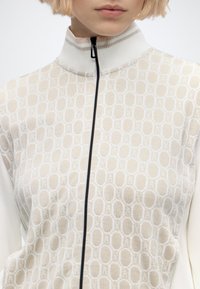 Gros plan d'une personne portant un haut beige à motifs avec un col montant zippé noir et des cheveux blonds courts.