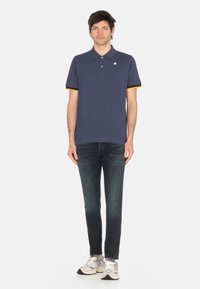Polo shirt blu scuro a maniche corte con dettagli gialli sui polsini e un logo bianco. Abbinato a jeans scuri e scarpe sportive.