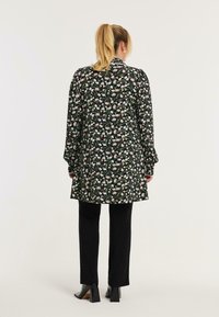 Langärmliges schwarzes Kleid mit Blumenmuster und Puffärmeln, über einer schwarzen, taillierten Hose, kombiniert mit knöchelhohen Stiefeletten mit Absatz.