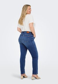 Denim jeans in dark blue with a smooth texture, high waist, back pockets, and a brown leather patch at the waistband. Paired with a white top.