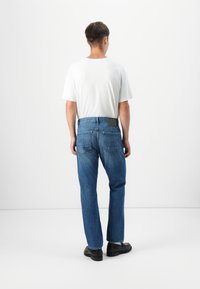 Jeans de denim azul con un corte recto, que cuentan con bolsillos traseros y un parche negro en la cinturilla, combinados con una camiseta blanca lisa.