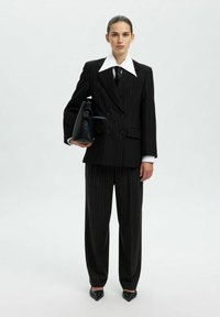 Črn pinstripe kostim z velikim belim ovratnikom, širokimi klapnicami in krojenimi hlačami. Dopolnjen z črno strukturirano torbico in špicastimi čevlji.