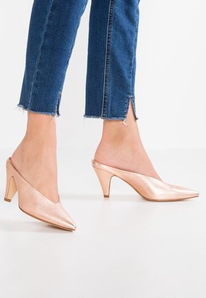 Metalické růžově zlaté mule s špičatou špičkou a 7 cm podpatkem, s elegantním designem a hladkým povrchem. Nosené s džínovými zkrácenými kalhotami.