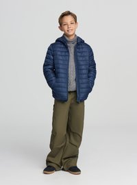 Giacca imbottita blu navy con cappuccio, indossata sopra un maglione grigio a trecce. Abbinata a pantaloni verde oliva a gamba larga e scarpe scure.