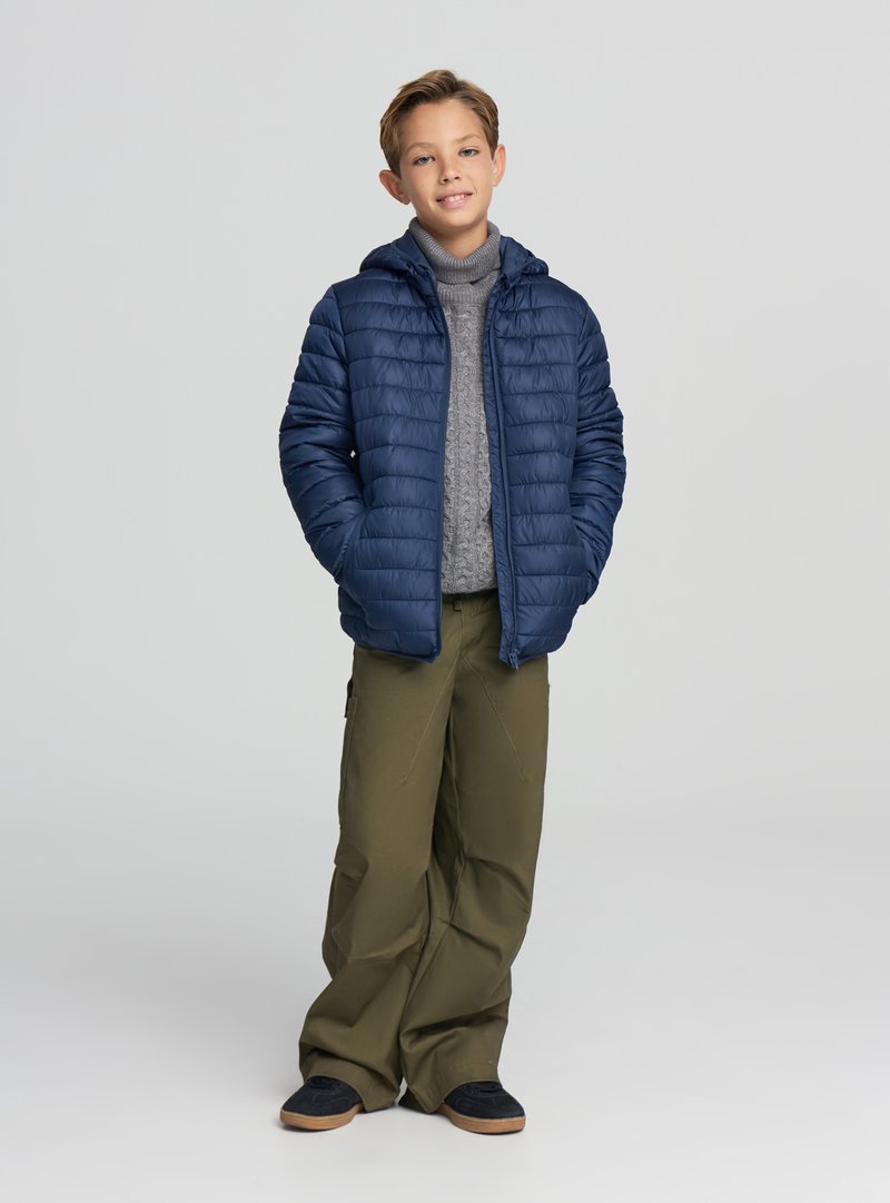 Giacca imbottita blu navy con cappuccio, indossata sopra un maglione grigio a trecce. Abbinata a pantaloni verde oliva a gamba larga e scarpe scure.