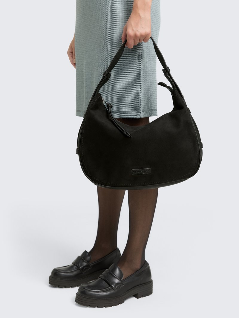 Černá semišová kabelka ve tvaru půlměsíce, s nastavitelným popruhem a zipovým uzávěrem. Umístěná na pozadí texturovaných světle modrých šatů a tmavých bot.