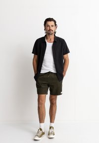 Camisa negra de manga corta, top blanco, pantalones cortos oliva, zapatillas deportivas casuales, pose de pie, entorno minimalista, manos en los bolsillos.