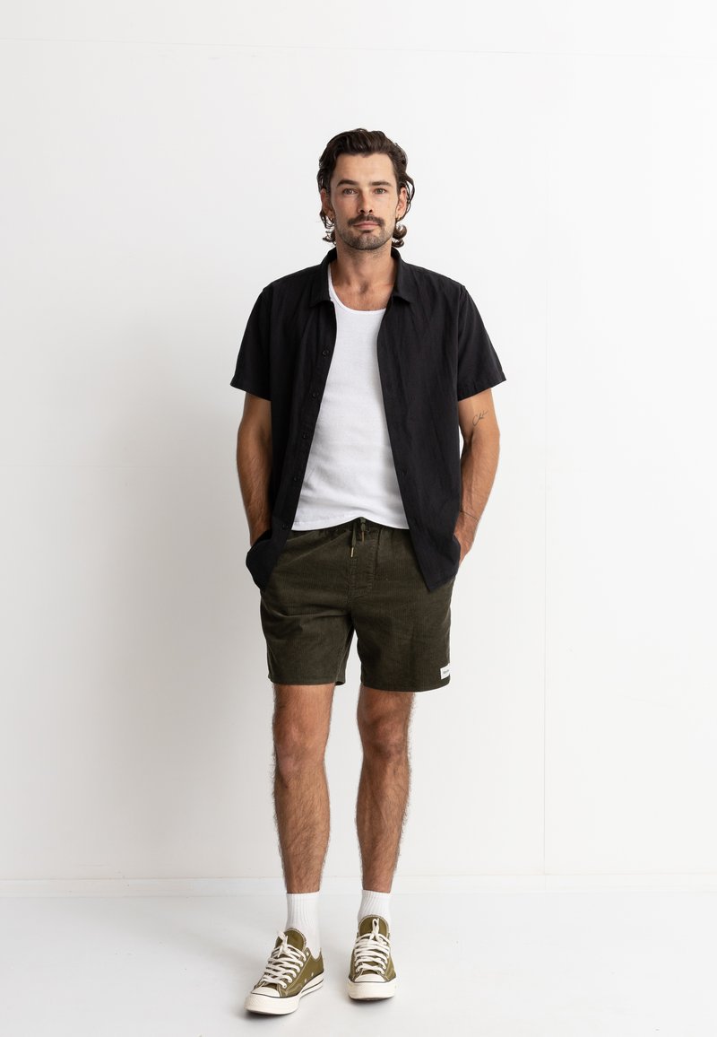 Camisa negra de manga corta, top blanco, pantalones cortos oliva, zapatillas deportivas casuales, pose de pie, entorno minimalista, manos en los bolsillos.