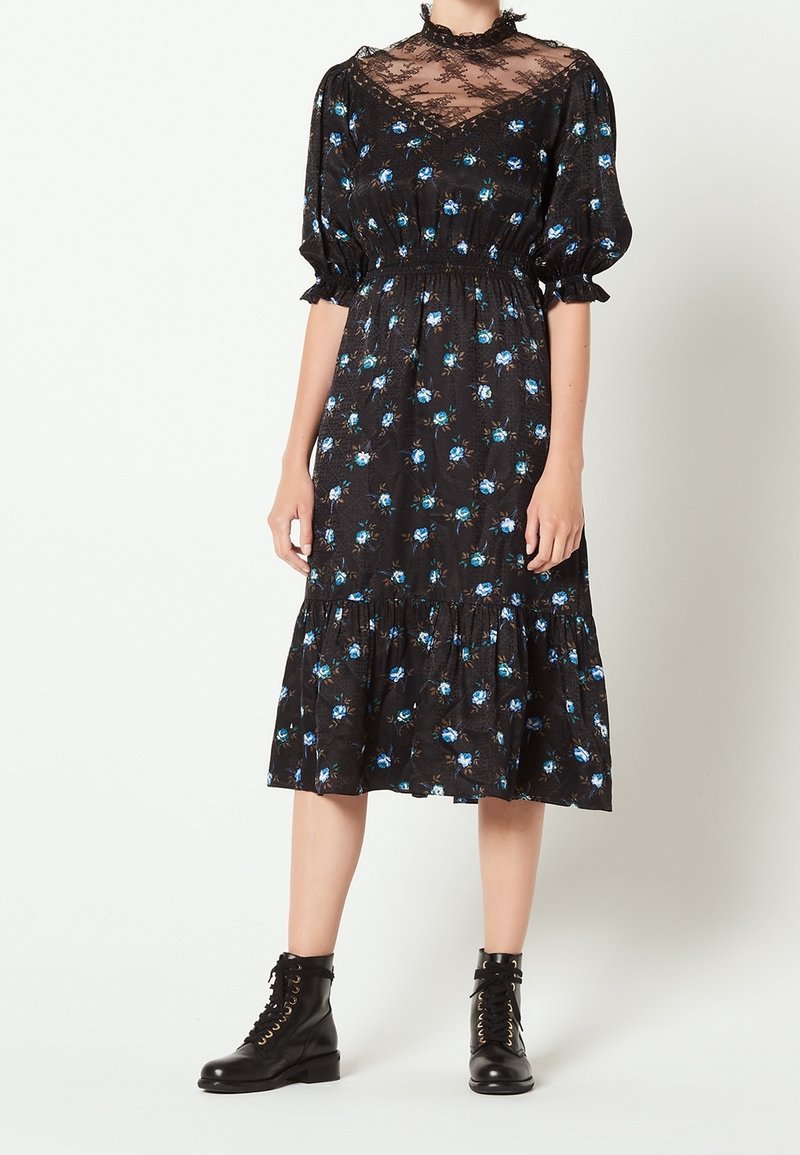Robe midi noire à imprimé floral bleu, col haut en dentelle transparente, manches bouffantes, ourlet à volants, associée à des bottines noires à lacets.