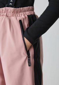 Rožinės ir juodos sportinės kelnės su raišteliu juosmenyje, šoninėmis užtrauktukų kišenėmis ir lygiu, matinio paviršiaus tekstūra. Etiketėje yra užrašas „BAKER“ balta spalva.