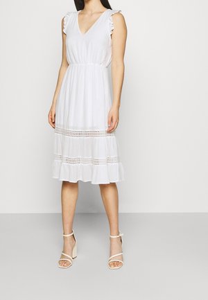 Femme portant une robe midi blanche sans manches avec des panneaux en dentelle et des détails à volants, associée à des sandales blanches à talons hauts, debout devant un fond uni.