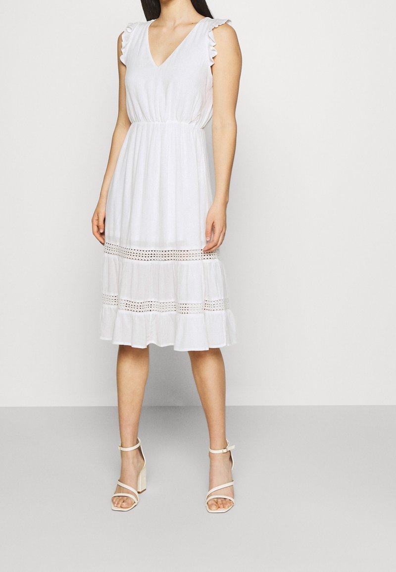 Femme portant une robe midi blanche sans manches avec des panneaux en dentelle et des détails à volants, associée à des sandales blanches à talons hauts, debout devant un fond uni.