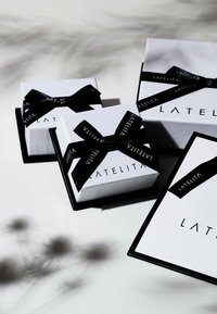 Tre scatole regalo bianche con nastro nero, con un logo. La scatola più grande è rettangolare, mentre le scatole più piccole sono quadrate e presentano dettagli di design simili.