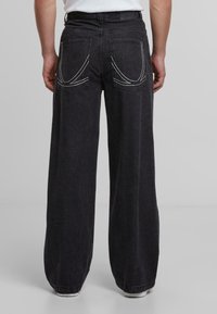 Pantalones de mezclilla negros de pierna ancha con costuras blancas, que cuentan con dos bolsillos traseros con costuras curvadas. La textura es suave con un ligero brillo.