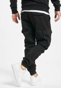Pantalon cargo noir avec poignets élastiques, doté de multiples poches et d'une coupe décontractée, porté avec des chaussures de sport blanches.