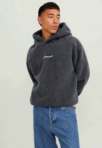 Jovem a usar um hoodie de fleece cinza escuro com o texto "originals studio", combinado com jeans azuis, olhando de lado com as mãos atrás das costas.