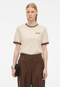T-shirt beige a maniche corte con un col ribattuto marrone scuro, caratterizzato da un logo stampato di colore marrone scuro. Abbinato a pantaloni marroni a vita alta.