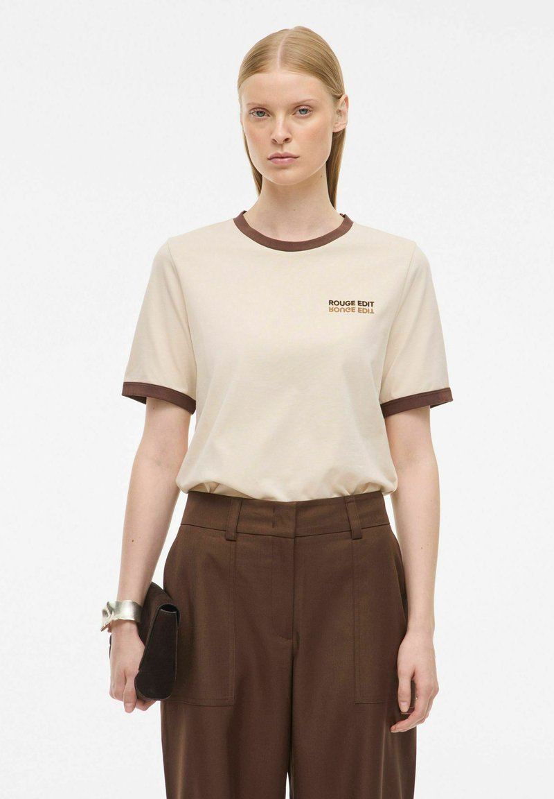 T-shirt beige a maniche corte con un col ribattuto marrone scuro, caratterizzato da un logo stampato di colore marrone scuro. Abbinato a pantaloni marroni a vita alta.
