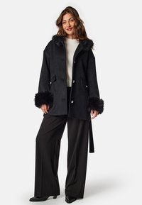 Black wool coat with plush fur accents on the cuffs and collar, featuring a straight cut, clasp buttons, and side pockets. Worn over a white sweater.