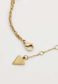 Bracciale in catena d'oro con un ciondolo triangolare inciso con la scritta "GUESS" e un gancio a moschettone per una chiusura sicura.