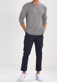 Mężczyzna w koszuli z długim rękawem w czarno-białe paski, ciemnych granatowych spodniach i białych sneakersach z czarnymi logo Swoosh, z rękami w kieszeniach.