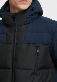 Chaqueta acolchada con capucha, paneles en contraste de azul oscuro y negro, textura suave, cremallera frontal y un bolsillo con cremallera en el pecho.