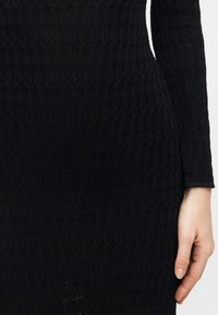 Κοντινό πλάνο ενός ατόμου που φοράει μαύρο, μακρυμάνικο φόρεμα πλεκτό με υφή και λεπτομερή μοτίβα, με το χέρι του να ακουμπά δίπλα στο σώμα.